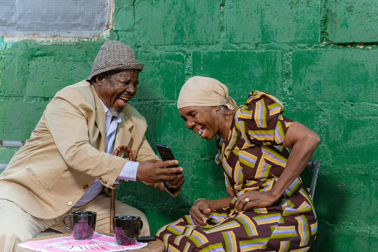 An older couple laughing together over a phone, he in a jacket and hat, she in a printed dress and headscarf, against a green wall
